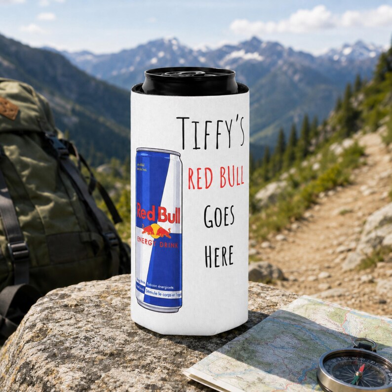 May include: A white can cooler with the text "TIFFY'S RED BULL GOES HERE" in black and red. A Red Bull energy drink can is visible on the cooler. The background shows a mountain range and a hiking trail.