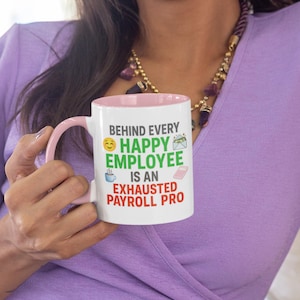 Peut inclure: Mug en céramique blanche avec intérieur et anse roses. Le mug affiche le texte "BEHIND EVERY HAPPY EMPLOYEE IS AN EXHAUSTED PAYROLL PRO" en noir, vert et rouge, ainsi que divers emojis. Le mug est tenu par une personne portant une chemise violette.
