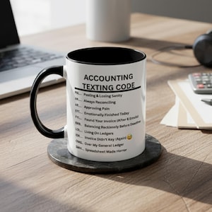 May include: White ceramic mug with a black interior and handle, featuring the text "ACCOUNTING TEXTING CODE" and accounting acronyms. The mug sits on a dark gray coaster. A laptop and other office supplies are in the background.