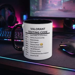 Può includere: Tazza in ceramica bianca con interno e manico neri. La tazza presenta il testo "VALORANT TEXTING CODE" e un elenco di abbreviazioni di gioco. La tazza è su una superficie nera con una tastiera e un mouse sullo sfondo.