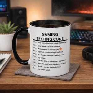 Può includere: Tazza in ceramica bianca con manico e bordo neri, con la scritta "GAMING TEXTING CODE" e acronimi di gioco con i loro significati. La tazza è appoggiata su un sottobicchiere grigio su una superficie di legno.