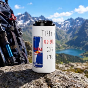 May include: A white beverage can cooler with the text "Tiffy's Red Bull Goes Here" and a Red Bull can graphic. The cooler is cylindrical and designed to hold a standard-sized can. The background shows a mountain range and a lake.