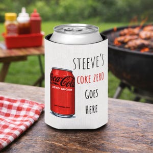 May include: A white can cooler with the text "STEEVE'S COKE ZERO GOES HERE" and a red Coca-Cola Zero can graphic. A silver soda can sits inside the cooler. The background includes a grill, condiments, and a checkered cloth.