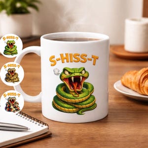 Puede incluir: Taza de cerámica blanca con el texto "S-HISS-T" y una ilustración de una serpiente de dibujos animados. La serpiente es verde con la boca abierta y la lengua roja. La taza está sobre una superficie de madera.
