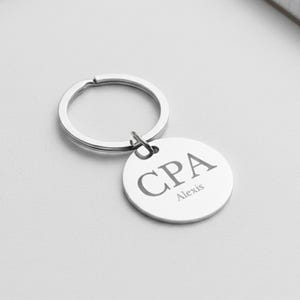 Può includere: Un portachiavi color argento con un ciondolo rotondo. Il ciondolo ha incise le lettere "CPA", con il nome "Alexis" sotto. Anche l'anello del portachiavi è argentato.