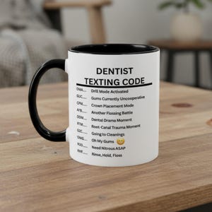 May include: A white ceramic mug with a black handle and rim. The mug features the text "DENTIST TEXTING CODE" and a list of dental-related abbreviations and their meanings, such as "DMA - Drill Mode Activated."