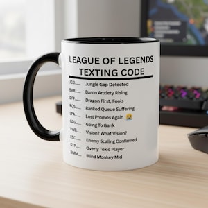 May include: White ceramic mug with a black handle and rim. The mug features the text "LEAGUE OF LEGENDS TEXTING CODE" and a list of gaming acronyms. The mug is on a light-colored surface.