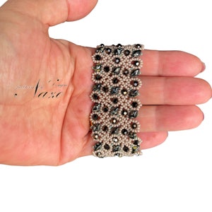 Op de afbeelding: Een kralenarmband met een geometrisch patroon in tinten zwart, grijs en roze. De armband is gemaakt met kleine kraaltjes en grotere facetgeslepen kralen.