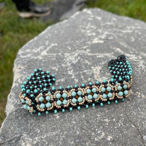 Könnte beinhalten: Ein türkisfarbenes und braunes Armband mit goldenen Akzenten. Das Armband ist aus kleinen, runden Perlen gefertigt und hat ein geometrisches Muster.