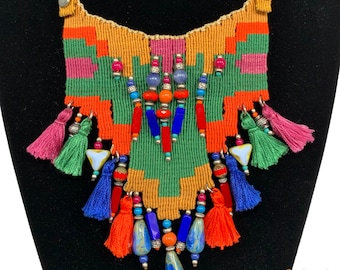 May include: A colorful woven necklace with tassels and beads. The necklace is made with green, orange, pink, red, blue, and white threads. It features a variety of beads, including turquoise, red, and blue.