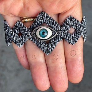 May include: A black and white macrame bracelet with a silver evil eye charm. The evil eye is blue and white.