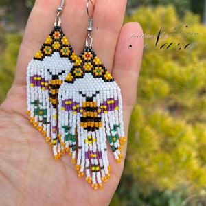 Pode incluir: Um par de brincos de contas brancas, amarelas, laranja, roxas, verdes e pretas com um design de abelha e franjas. Os brincos estão pendurados em ganchos prateados.