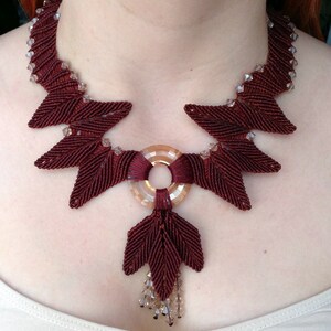May include: A maroon and brown necklace with a circular pendant and a dangling leaf design. The necklace is made of cord and features clear crystal beads.