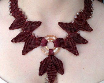 May include: A maroon and brown necklace with a circular pendant and a dangling leaf design. The necklace is made of cord and features clear crystal beads.