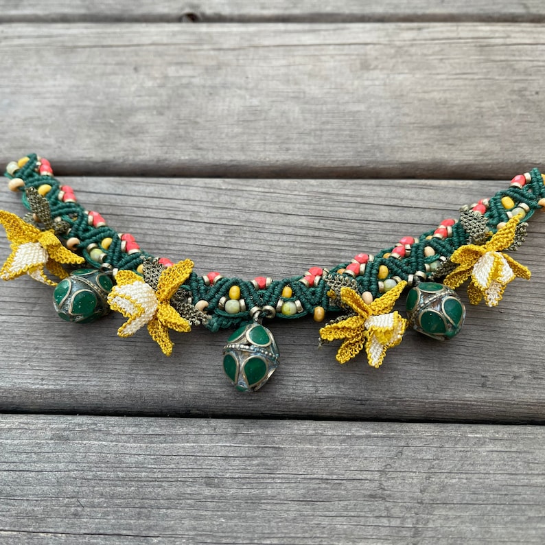 May include: A green and red macrame necklace with yellow crocheted flowers and green metal bells.