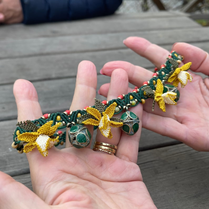 May include: A green, yellow, and red crocheted bracelet with small green and silver bells. The bracelet is decorated with yellow flower-shaped charms.