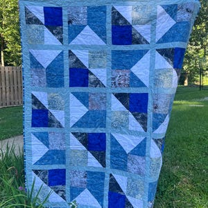 May include: A rectangular quilt featuring a grid of square blocks in various shades of blue, white, and grey. The design consists of alternating solid blue, white, and marbled patterned fabric triangles arranged in a geometric pinwheel-like pattern.
