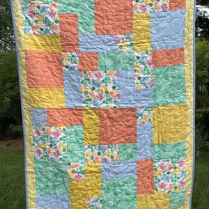 May include: A handmade quilt with a patchwork design. The quilt features squares of peach, yellow, light blue, and green fabric. Some squares have a floral pattern. The quilt is bordered with yellow and blue fabric.