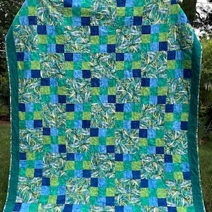 May include: A handmade quilt with a patchwork design. The quilt features a pattern of alternating squares in shades of blue and green, with a teal border. The squares are arranged in a grid pattern, creating a visually appealing textile.