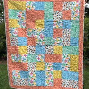May include: A handmade patchwork quilt with a variety of colorful squares. The quilt features squares of yellow, light blue, peach, and green. Some squares have a floral pattern. The quilt is trimmed with a peach border.