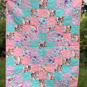 May include: A patchwork quilt with a pink border and backing. The quilt features squares of pink, light blue, and lavender fabric, some with floral patterns and others with a repeating doll design. The quilt is held up against a green background.