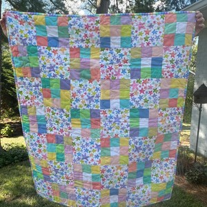 May include: A handmade quilt featuring a patchwork design with squares in pastel colors. The quilt has a white background with colorful star patterns. The squares are in shades of pink, yellow, green, and blue. The quilt is held up by a person.