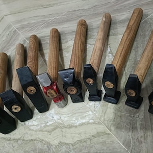 Puede incluir: Una colección de martillos con mangos de madera y cabezas negras o plateadas. Los martillos están dispuestos en fila, mostrando sus diferentes tamaños y diseños. Los mangos de madera tienen un acabado natural.