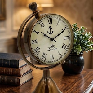 May include: A brass-toned table clock with a circular frame and a pedestal base. The off-white clock face features black Roman numerals, a central anchor icon, and the text London with a small crown graphic above it, set with black hands.