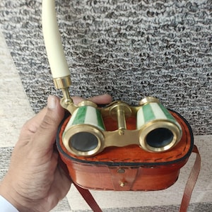 May include: Vintage opera glasses with a cream-colored handle and green and white striped accents. The brass-toned binoculars are in a brown leather case with a matching strap. A classic accessory for theater or travel.