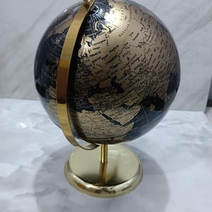 Puede incluir: Un globo terráqueo decorativo con una esfera negra y continentes y texto en oro metálico. El globo está montado sobre un soporte de metal de color dorado pulido con una base circular y un anillo meridiano vertical que sujeta la esfera.