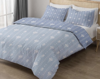 Puede incluir: Un juego de cama azul claro con funda nórdica y dos fundas de almohada. La ropa de cama presenta un patrón repetido de diseños florales blancos y rayas verticales. La cama tiene un cabecero de madera.