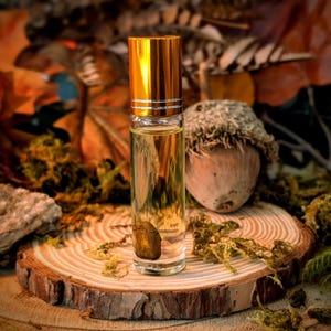 May include: A clear glass essential oil roller bottle with a gold cap, containing a small stone. The bottle sits on a wooden slice, with an acorn and moss in the background. The background features autumn leaves.