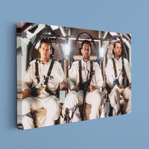 May include: A rectangular canvas print featuring a scene from the film Apollo 13, showing three astronauts in white flight suits and harnesses seated inside a spacecraft cockpit, with metallic structural elements visible in the background.