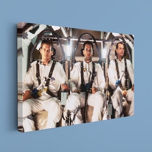 May include: A canvas print featuring three individuals in white flight suits seated in a cockpit. The image is a still from a film, with the subjects appearing to be astronauts or pilots. The canvas is mounted on a blue wall.