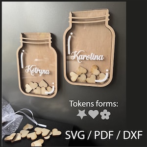 以下が含まれることがあります： 透明なアクリル製の前面を備えた、木製のメイソンジャー型の装飾ボックスが2つあります。「Kotryna」と「Karolina」という名前が白い文字で刻まれています。小さな木製のハート型のトークンがジャーの中にあります。「Tokens forms:」と「SVG / PDF / DXF」というテキストも表示されています。