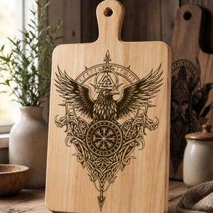 Peut inclure: Une planche à découper rectangulaire en bois clair avec une poignée et un trou pour la suspendre. La surface présente un motif gravé au laser représentant un corbeau aux ailes déployées, un symbole Valknut et une boussole Vegvisir.