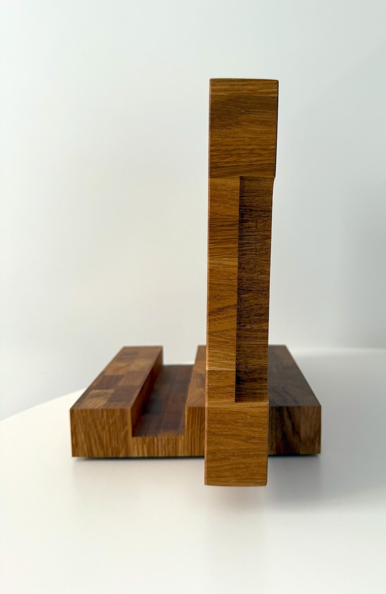 K&ouml;nnte beinhalten: Ein h&ouml;lzerner Telefonst&auml;nder mit geometrischem Design. Der St&auml;nder besteht aus hellem und dunklem braunem Holz mit einer rechteckigen Basis und einer vertikalen St&uuml;tze. Der St&auml;nder ist f&uuml;r ein Telefon oder Tablet konzipiert.