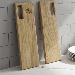 May include: Two light-colored wooden serving boards with a rectangular shape and a handle cutout. One board has a circular logo. The boards are displayed on a white countertop with a tiled background, suggesting a kitchen setting.