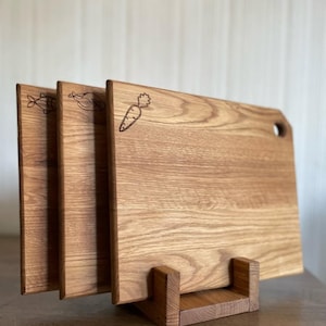 Könnte beinhalten: Drei Holzschneidebretter mit eingravierten Lebensmittelillustrationen: ein Fisch, ein Huhn und eine Karotte. Die Bretter werden in einem Holzständer präsentiert, der ihre natürliche Holzmaserung und rechteckige Form mit abgerundeten Ecken zeigt. Ein Brett hat ein Loch.