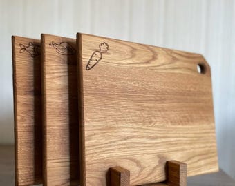 May include: Three wooden cutting boards with engraved food illustrations: a fish, a chicken, and a carrot. The boards are displayed in a wooden stand, showcasing their natural wood grain and rectangular shape with rounded corners. One board has a hole.
