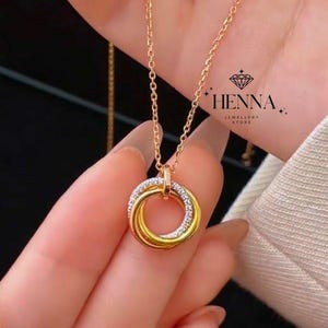 May include: A gold-coloured necklace with three intertwined rings, one set with small, clear crystals. The pendant is suspended from a fine chain. The image includes the text "HENNA JEWELLERY STORE".
