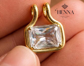 May include: A gold pendant featuring a rectangular clear gemstone set in a smooth metal frame with two top loops for attachment. The pendant includes the text HENNA JEWELLERY STORE with a diamond icon printed in the upper right corner of the image.