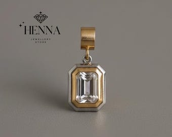 May include: A rectangular pendant with a clear, emerald-cut gemstone. The pendant has a gold-colored frame around the gemstone, with a silver-colored outer frame. The top loop is gold-colored. The logo "HENNA JEWELLERY STORE" is in the upper left corner.