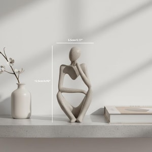 Op de afbeelding: Een neutrale sculptuur van een zittende figuur met een hand op de kin, 12,5 cm hoog en 5,5 cm breed. Het minimalistische ontwerp wordt getoond op een plank met een vaas en een boek, wat een serene, artistieke weergave creëert.