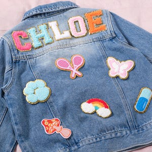 Op de afbeelding: Een lichtblauwe spijkerjas met de naam "CHLOE" geborduurd op de rug in kleurrijke, getextureerde letters. De jas is versierd met patches van een tennisracket, vlinder, wolk, regenboog, paddenstoel en een capsule.