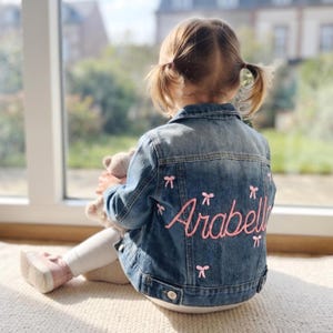 May include: A blue denim jacket featuring the name Arabella embroidered in pink script on the back, accented with small pink embroidered bows scattered across the fabric. The jacket has a classic collar, buttoned cuffs, and metal buttons.