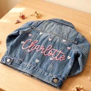 May include: A blue denim jacket featuring the name Charlotte embroidered in pink yarn across the back. The jacket is decorated with small, scattered embroidered hearts in shades of pink and white, and includes metal buttons on the cuffs and hem.