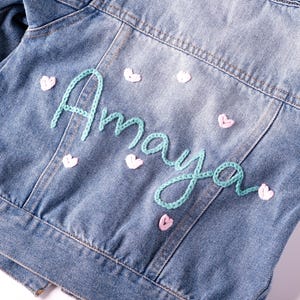 May include: Pale blue denim jacket featuring the name "Amaya" embroidered in turquoise script on the back. Small pink heart-shaped embellishments are scattered around the name. A classic button-down jacket.