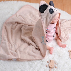 May include: A baby wrapped in a soft, pale pink hooded towel with panda ears and a matching blanket. The towel is personalised with the name "Jacob" in blue script. Wooden toys are nearby.
