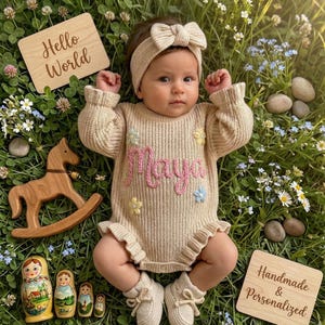 May include: A baby in a beige knitted romper with the name "Maya" embroidered in pink, lying on grass with flowers and wooden toys. A wooden sign says "Hello World". The romper has a bow and is paired with matching booties.